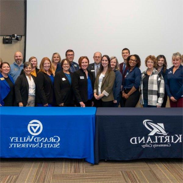 large group of people standing in two rows behind 2 tables draped with banners for GVSU and Kirtland Community College